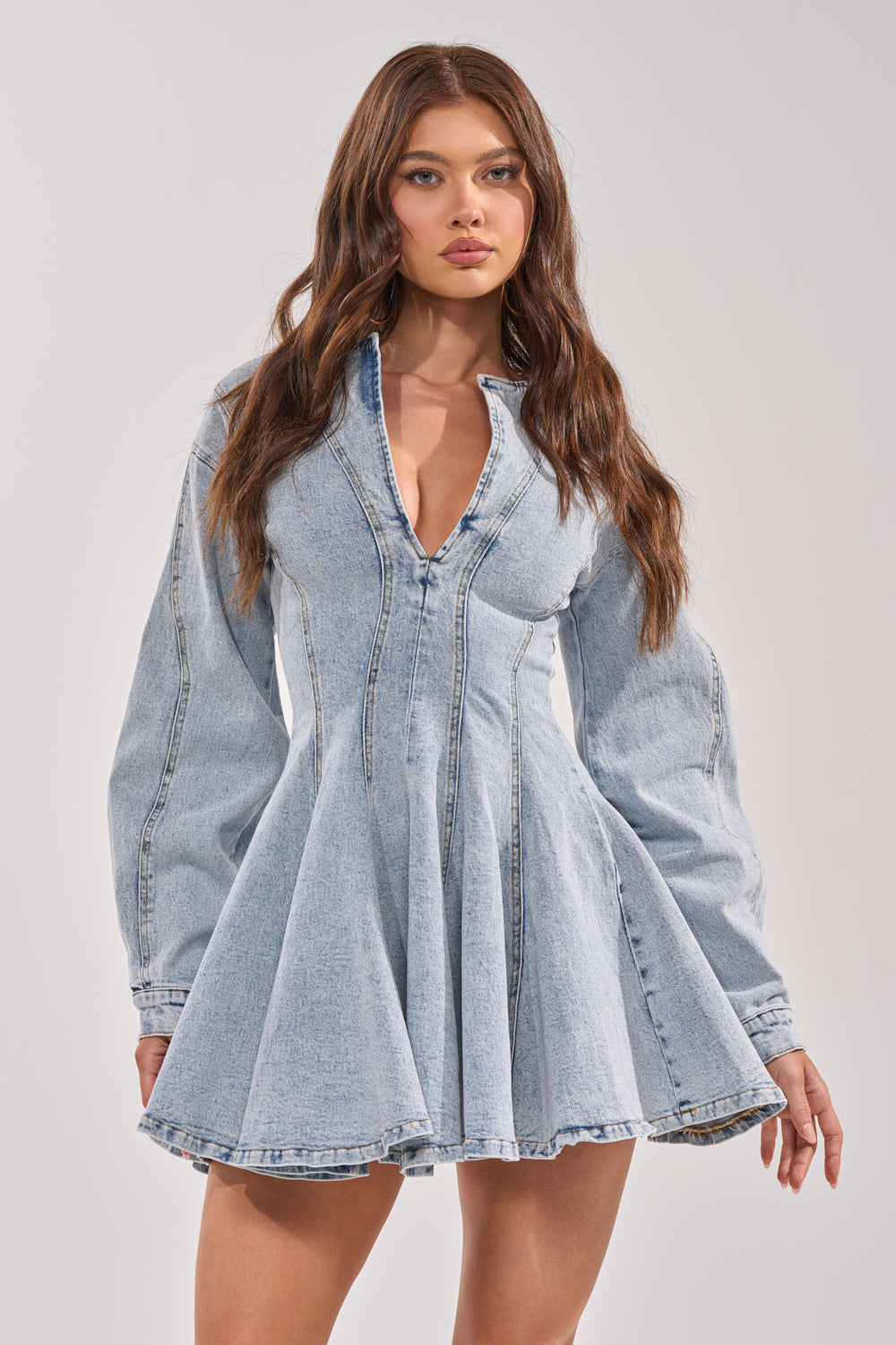 A woman with long brown hair models the ALL ABOUT MY BUSINESS DENIM MINI DRESS, a light wash fit and flare mini dress with long sleeves and a deep V-neckline, standing against a neutral background.