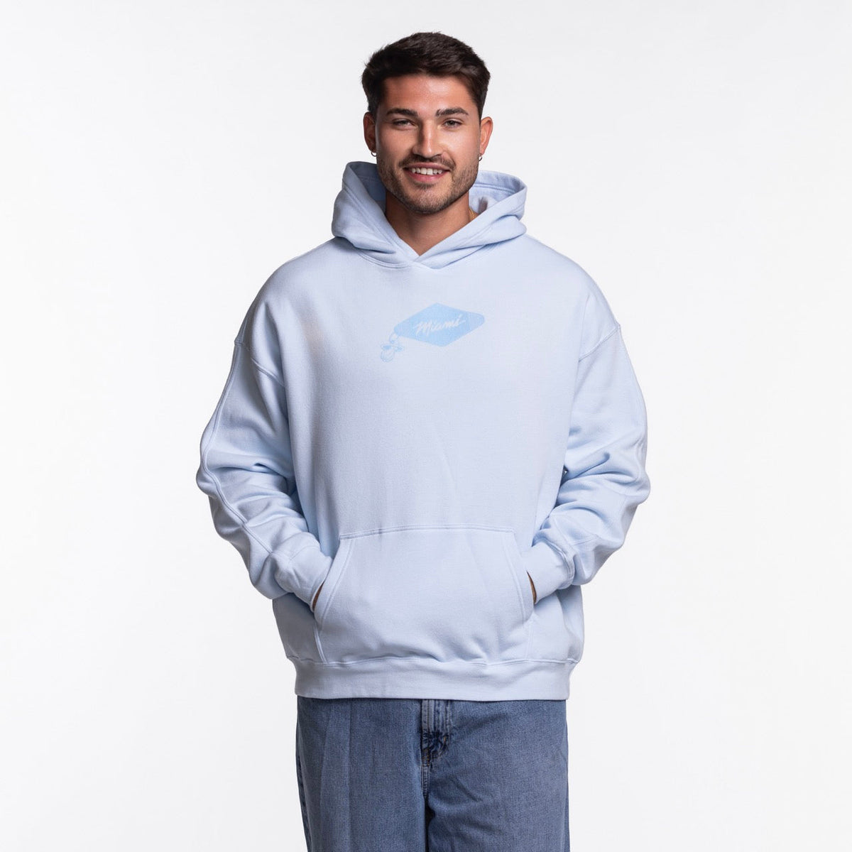 A man stands facing forward, smiling with hands in the pocket of a light blue Court Culture Original Vice Nights "Drive-In" Oversized Unisex Hoodie, featuring a small chest graphic. He pairs it with blue jeans against a white background.