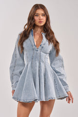 A woman with long brown hair models the ALL ABOUT MY BUSINESS DENIM MINI DRESS, a light wash fit and flare mini dress with long sleeves and a deep V-neckline, standing against a neutral background.
